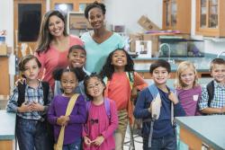 A Black and a white co-teacher stand with their group of young children in the classroom.