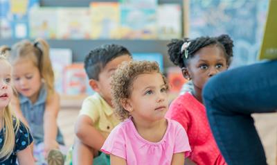 Children sit on the floor in a group as their teacher reads a story